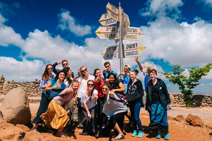 A group of travelers posing at a signpost that points to Damascus, Tiberias and other places